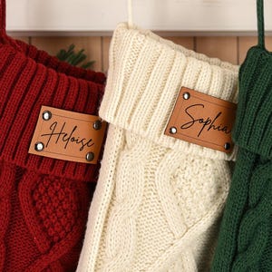 Op de afbeelding: Drie kabelgebreide kerstsokken in rood, crème en groen hangen aan een schoorsteenmantel. Elke sok heeft een leren naamlabel met een naam in cursief. De sokken zijn versierd met kerstversiering.