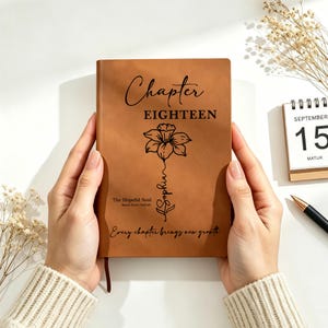 May include: A brown leather journal with the words "Chapter EIGHTEEN" and a floral illustration. The journal also features the text "The Hopeful Soul" and "Every chapter brings new growth". A calendar showing September 15th is in the background.