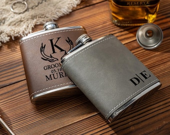 May include: Two personalized flasks, one brown and one gray, with silver accents. The brown flask has the letter "K" and the text "GROOM 05.16 MUR" engraved. The gray flask has the initials "DE". A bottle of whiskey is in the background.