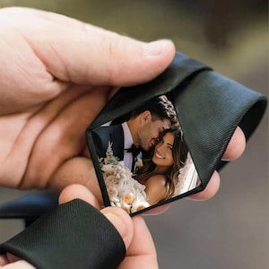 Könnte beinhalten: Eine schwarze Krawatte mit einem versteckten Foto eines lächelnden Paares. Das Foto zeigt einen Mann im Anzug und eine Frau im Brautkleid mit einem Blumenstrauß. Die Krawatte wird von einer Hand gehalten.