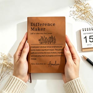 Op de afbeelding: Een bruin leren notitieboek met de woorden "Difference Maker" en de definitie erop gedrukt. Het notitieboek bevat ook de naam "Amelia". Een kalender met 15 september staat op de achtergrond.