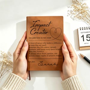 Könnte beinhalten: Ein Notizbuch mit beigem Ledereinband, bedruckt mit dem schwarzen Text Impact Creator, einer phonetischen Aussprache, einer Definition, einem gepunkteten Herzumriss und dem Namen Sarah, gehalten von zwei Händen in cremefarbenen Ärmeln.