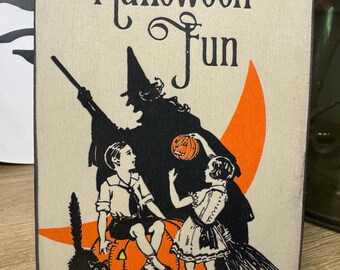 May include: Wooden Halloween sign with the words "Halloween Fun" in black script. The sign features a witch silhouette, children, a carved pumpkin, and a black cat. The text "The Big Book of All Hallows' Eve" is at the bottom.