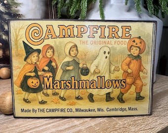 May include: Wooden sign with a vintage illustration featuring children in Halloween costumes. The sign reads "Campfire Marshmallows" with text "The Original Food." The sign is made by The Campfire Co. in Milwaukee, Wisconsin and Cambridge, Massachusetts.