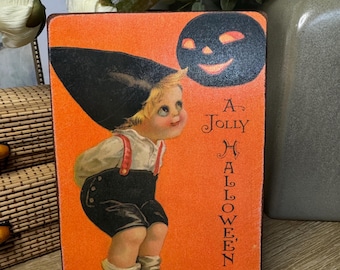 May include: Vintage Halloween-themed wooden block sign featuring a child in a black hat and shorts, looking up at a smiling jack-o'-lantern. The sign has an orange background with the words "A Jolly Halloween" in vertical text.