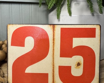 May include: A rectangular wooden sign with the number 25 in bold, bright red numerals. The numbers are set against a cream-colored background with a distressed, vintage aesthetic. The sign is likely for home decor.