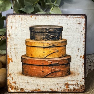 May include: A square wooden sign with a distressed white background and a brown border. The sign features a stack of three antique wooden boxes in shades of brown, yellow, and black. The boxes have distinctive wooden closures.