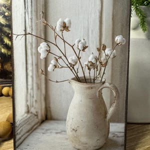 May include: A rustic wooden sign featuring a still life of cotton stems in a cream-colored pitcher. The pitcher has a distressed finish, and the background shows a window with a soft, neutral color palette. The overall aesthetic is farmhouse style.