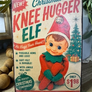 May include: A vintage-style Christmas sign featuring a red-suited elf with a green collar and a jingle bell hat. The sign reads "Christmas Knee Hugger Elf" with text that says "He Hugs Your Heart!" and "Makes Every Season Merry!"