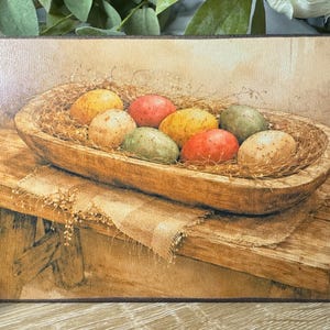 May include: A rectangular wooden sign featuring a still life painting of colorful Easter eggs in a wooden bowl. The eggs are in shades of yellow, red, green, and tan, nestled in straw. The bowl sits on a rustic wooden table.