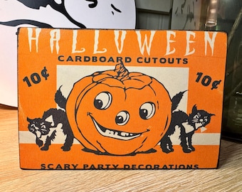 May include: Vintage Halloween cardboard cutout with an orange background. The cutout features a smiling jack-o'-lantern, two black cats, and the text "HALLOWEEN CARDBOARD CUTOUTS" and "SCARY PARTY DECORATIONS".