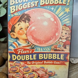 May include: Vintage-style sign featuring a boy blowing a large pink bubble, with a girl holding packs of gum. The sign reads "Blow the Biggest Bubble!" and "Fleer's Double Bubble - The Original Bubble Gum!" with images of gum.
