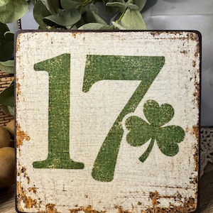 May include: Square wooden sign with a distressed white background and green lettering. The number "17" is prominently displayed, accompanied by a green shamrock. The sign has a rustic, vintage aesthetic with visible wear and tear.