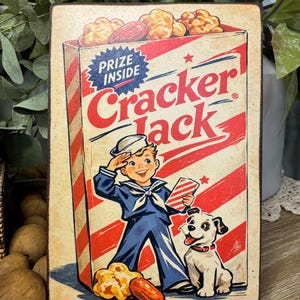May include: Vintage-style wooden sign featuring a Cracker Jack box design. The box illustration includes a boy in a sailor suit, a dog, and the words "Prize Inside" and "Cracker Jack" in red and blue.