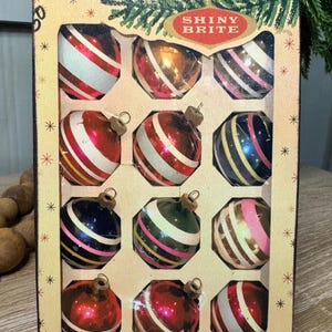 May include: Vintage Shiny Brite glass Christmas tree ornaments in a box. The ornaments are red, white, gold, and blue with striped designs. The box is tan with the words "Shiny Brite" and "American Made Glass Christmas Tree Ornaments".