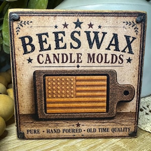 May include: A rustic wooden sign with the words "BEESWAX CANDLE MOLDS" in black lettering. Below is a carved wooden American flag. The sign also includes the phrases "PURE", "HAND POURED", and "OLD TIME QUALITY".
