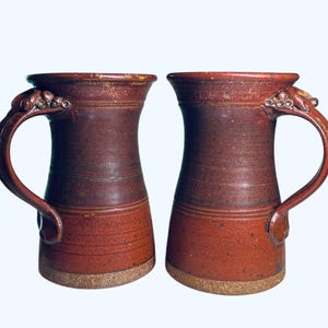 May include: Two handcrafted ceramic mugs with a rustic, textured appearance. The mugs are a gradient of reddish-brown hues, with a lighter base and darker upper portion. Each mug features a curved handle and decorative detailing near the rim.