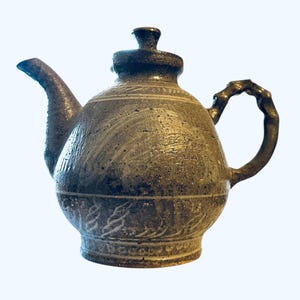 May include: A handcrafted ceramic teapot with a dark, textured finish. The teapot features a curved spout, a loop handle, and a lid with a small knob. Decorative etched patterns adorn the body of the teapot, adding visual interest.