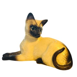 May include: A ceramic Siamese cat figurine in a resting pose. The cat has a cream-colored body with dark brown markings on its face, ears, paws, and tail. The figurine is glossy and detailed.