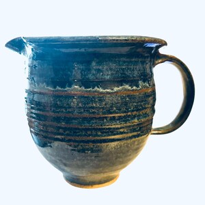 May include: A handmade ceramic pitcher with a dark blue and brown glaze. The pitcher has a spout, a handle, and horizontal textured lines around the body. The base is a lighter tan color.