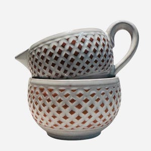 May include: A white ceramic creamer and bowl set. The creamer has a handle and spout, and is stacked on top of the bowl. Both pieces feature a lattice design with brown accents.