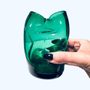 May include: A green glass tumbler with a unique, asymmetrical shape. The glass has a faceted design and a glossy finish. The tumbler is held by a hand with dark nail polish. The background is a solid white.