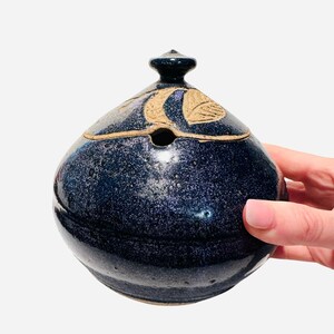 May include: A small, round, blue ceramic jar with a lid. The lid has a small hole in the center and is decorated with a brown leaf design.
