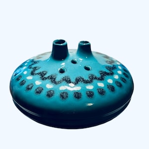 May include: A turquoise ceramic decorative piece with two spouts and a series of small holes. The piece is adorned with a pattern of black and white dots and a zigzag design. The overall shape is round and low-profile.
