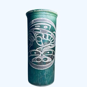May include: A cylindrical, teal-colored ceramic vase with a detailed, etched design. The design features a stylized, symmetrical motif in silver, possibly depicting a bird or other creature. The vase has a slightly textured surface.