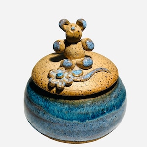 May include: A handcrafted ceramic jar with a lid, featuring a charming mouse figurine on top. The jar is a vibrant blue with a speckled tan lid, adorned with a blue flower. This decorative piece is ideal for storing small items.