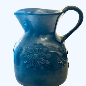 May include: A blue ceramic pitcher with a curved handle and a spout. The pitcher features a raised floral design on the front and leaves on the side. The pitcher is a decorative item.