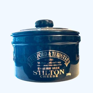 May include: A dark blue Stilton cheese crock with a lid. The crock has gold lettering that reads "Tuxford & Tebbutt" and "English Blue Stilton Cheese." The crock contains 250g of cheese.