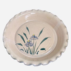 May include: A cream-colored ceramic plate with a ruffled edge. The center of the plate features a hand-painted design of blue irises with green leaves and stems. The plate has a rustic, handmade appearance.