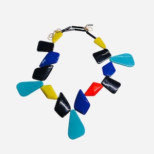 May include: A colorful necklace featuring geometric shapes in black, blue, yellow, red, and turquoise. The necklace has a black cord and gold-colored clasp. The shapes are arranged in a symmetrical pattern, creating a bold and modern accessory.