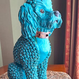 Op de afbeelding: Turquoise keramieken poedelbeeld met een getextureerde vacht en gedetailleerde kenmerken. De hond zit, met een bronskleurige halsband. Het beeld staat op een oppervlak met patronen, met een gestreepte achtergrond en een rood gordijn.