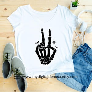 May include: White V-neck t-shirt featuring a black skeleton hand making a peace sign. The design includes bats and a spider. The text "http://www.mydigitaldenusa.etsy.com" is printed at the bottom of the shirt.