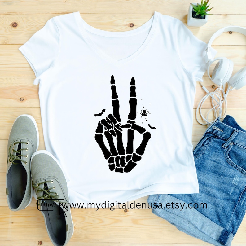May include: White V-neck t-shirt featuring a black skeleton hand making a peace sign. The design includes bats and a spider. The text "http://www.mydigitaldenusa.etsy.com" is printed at the bottom of the shirt.