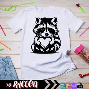 Pu&ograve; includere: Una maglietta bianca con un'illustrazione in bianco e nero di un procione che tiene un cuore. Il testo "30 RACCOON" &egrave; nella parte inferiore dell'immagine.
