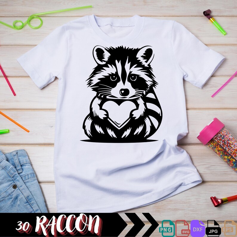 Pu&ograve; includere: Una maglietta bianca con un'illustrazione in bianco e nero di un procione che tiene un cuore. Il testo "30 RACCOON" &egrave; nella parte inferiore dell'immagine.