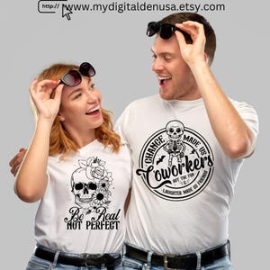 May include: Two people wearing white t-shirts with graphic designs. One shirt features a skull with flowers and the text "Be Real Not Perfect." The other shirt has a skeleton and the text "Coworkers but the fun, laughter made us friends."