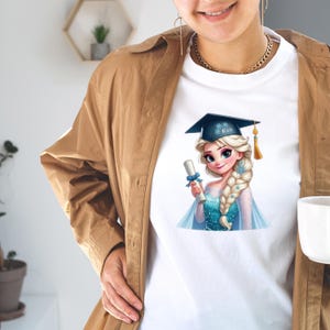 May include: White t-shirt featuring a cartoon illustration of a blonde woman wearing a graduation cap and holding a diploma. The woman is wearing a blue dress and has a long braid. The graduation cap has the name "Elsa" on it.
