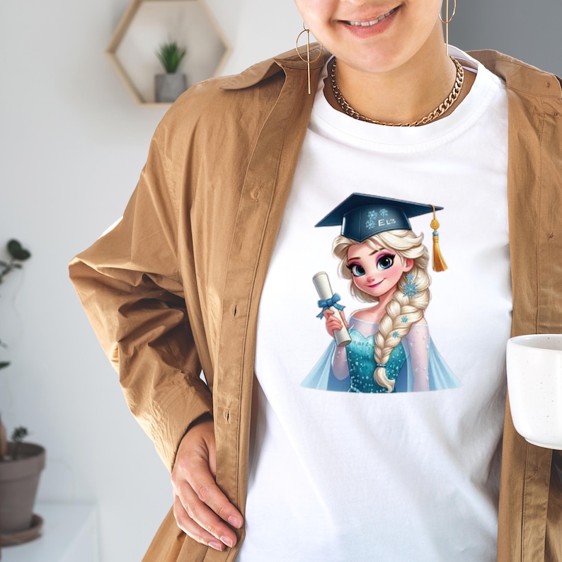May include: White t-shirt featuring a cartoon illustration of a blonde woman wearing a graduation cap and holding a diploma. The woman is wearing a blue dress and has a long braid. The graduation cap has the name "Elsa" on it.