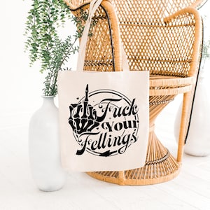 May include: Beige tote bag with black graphic text that reads "Fuck Your Feelings" with a skeleton hand giving the middle finger. The bag has a simple design and is made of a canvas-like material. The bag is hanging on a wicker chair.