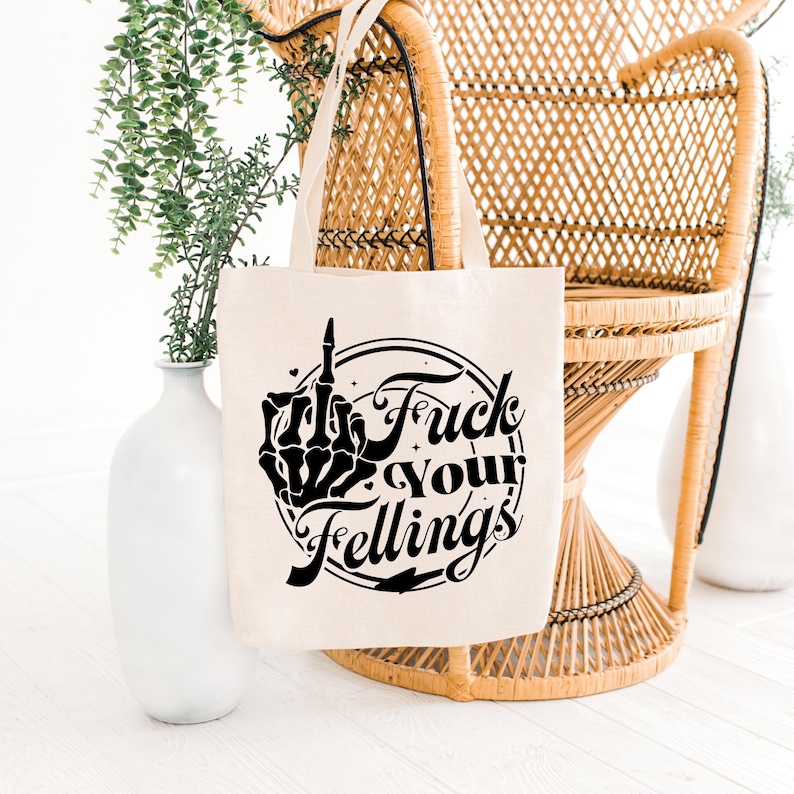 May include: Beige tote bag with black graphic text that reads "Fuck Your Feelings" with a skeleton hand giving the middle finger. The bag has a simple design and is made of a canvas-like material. The bag is hanging on a wicker chair.