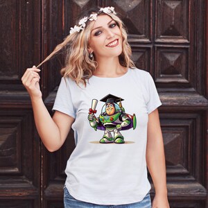 May include: Light blue t-shirt featuring a cartoon illustration of Buzz Lightyear in a graduation cap, holding a diploma. The character is depicted in his signature green, white, and purple spacesuit. The shirt is a crew neck style.