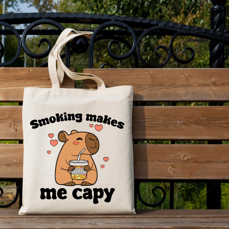 Pu&ograve; includere: Borsa tote beige con la scritta nera "Smoking makes me capy". La borsa presenta un disegno di un capibara che tiene una bevanda, circondato da cuori rosa. La borsa ha due manici.