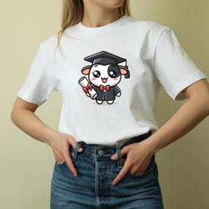 May include: White t-shirt featuring a cartoon cow wearing a graduation cap and gown, holding a diploma. The cow has a black and white face, a red bow tie, and is smiling. The shirt is plain white.