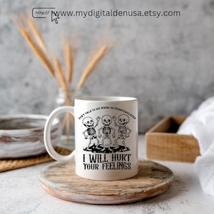 May include: White ceramic mug with a black graphic of three skeletons and the text "DON'T TALK TO ME WHEN I'M OVERSTIMULATED" and "I WILL HURT YOUR FEELINGS". The mug is on a wooden coaster.