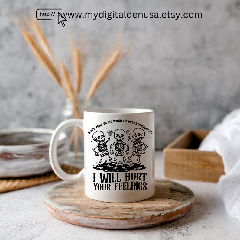 May include: White ceramic mug with a black graphic of three skeletons and the text "DON'T TALK TO ME WHEN I'M OVERSTIMULATED" and "I WILL HURT YOUR FEELINGS". The mug is on a wooden coaster.