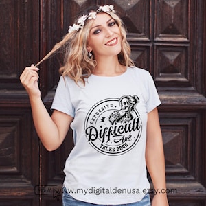 May include: A light blue t-shirt with a black and white graphic. The graphic is a circular design with the words "Expensive, Difficult, And Talks Back" surrounding a skeleton woman holding a can. The Etsy shop URL is visible.